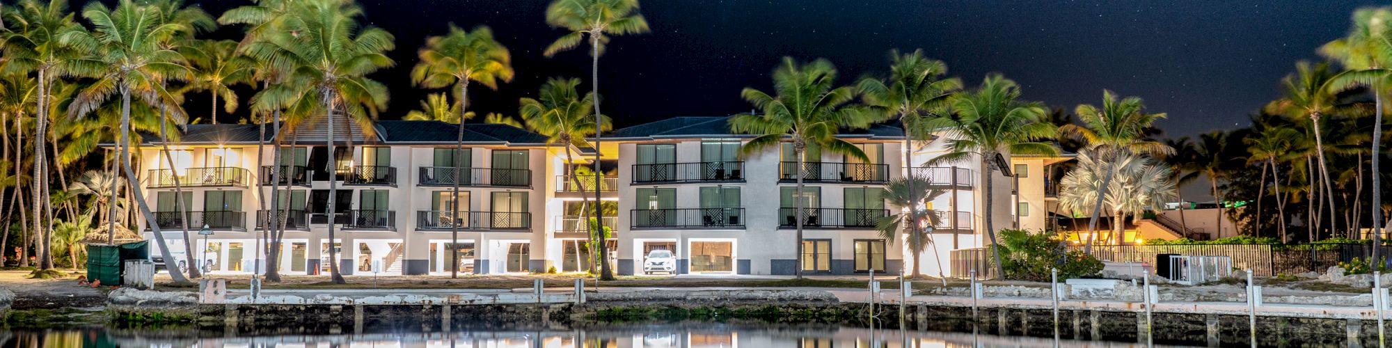 A night view of a waterfront building with lights on, surrounded by palm trees, reflecting in a calm body of water under a starry sky.