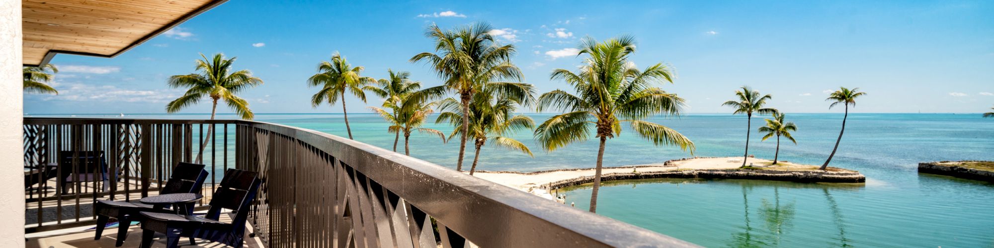 A balcony with lounge chairs, overlooking a serene beach scene with palm trees and calm ocean waters under a clear, bright blue sky.