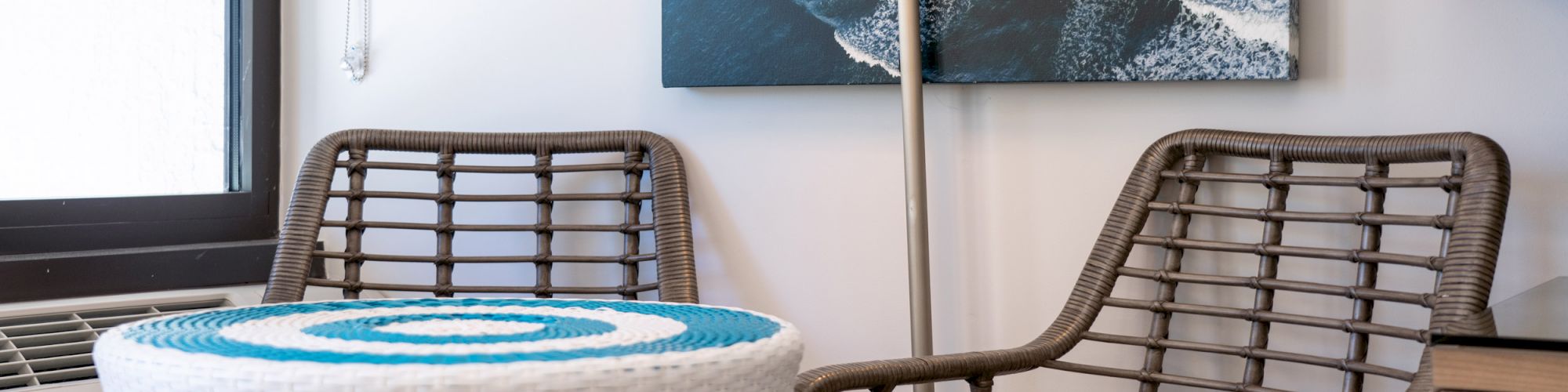 The image shows two wicker chairs, a blue and white striped wicker table, a standing lamp, and wall art featuring waves next to a window.