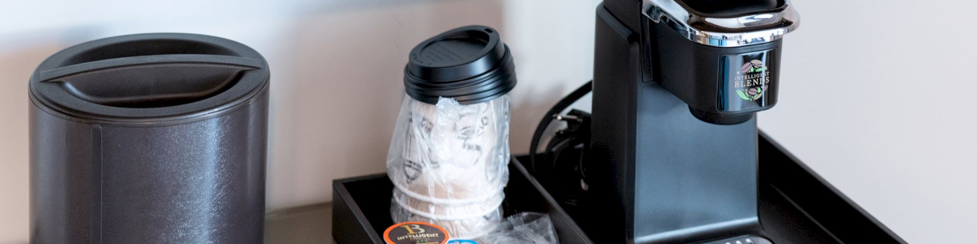 The image shows a coffee station with a small coffee machine, coffee pods, a wrapped cup, and a trash can on a countertop.