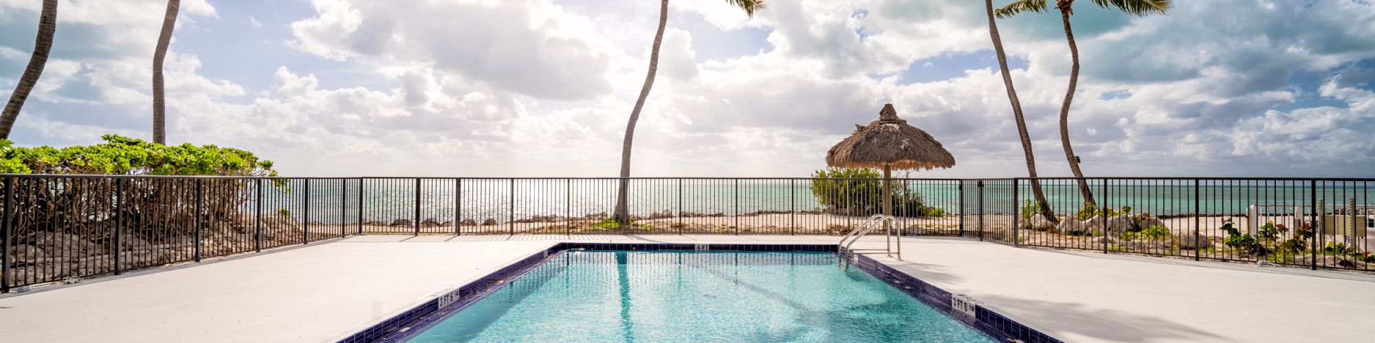 A serene swimming pool surrounded by palm trees, overlooking the ocean with a partly cloudy sky and sun peeking through clouds in the background.