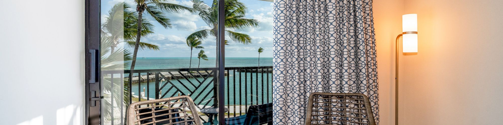 A room with two wicker chairs, a small table, a floor lamp, and a window showing a balcony with a view of the ocean and palm trees.