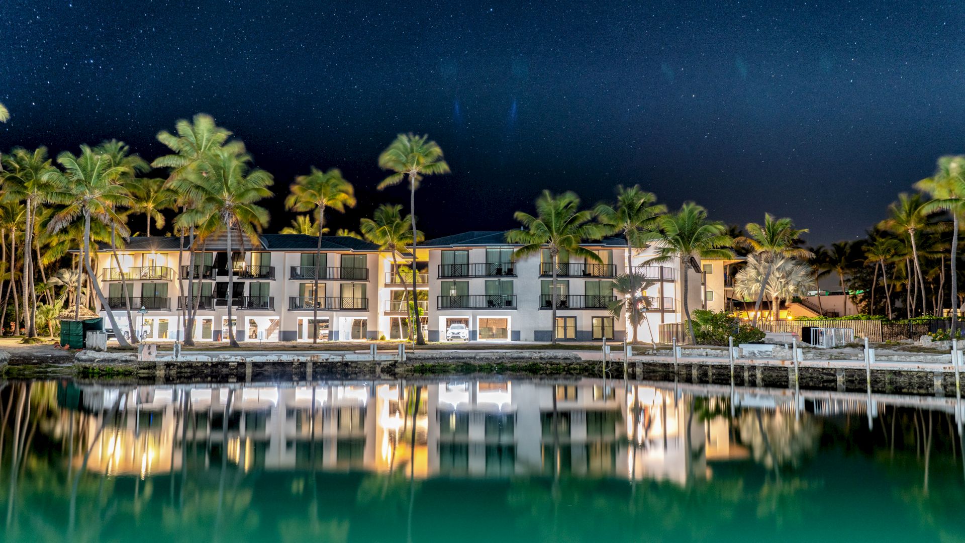 The image shows a waterfront hotel or apartment complex at night, surrounded by palm trees, with its reflection visible in the calm water.