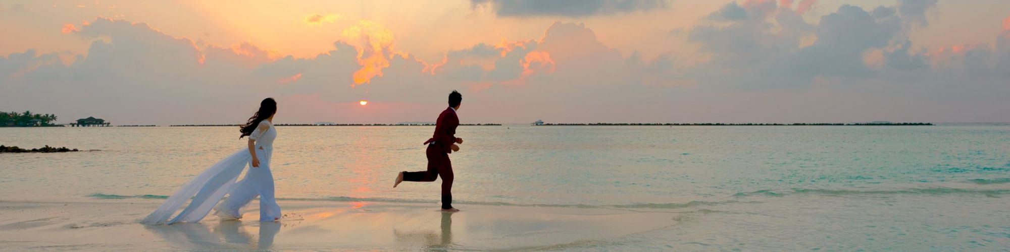 A woman in a white dress and a man in dark clothing are playfully running on a shallow beach during a beautiful sunset.