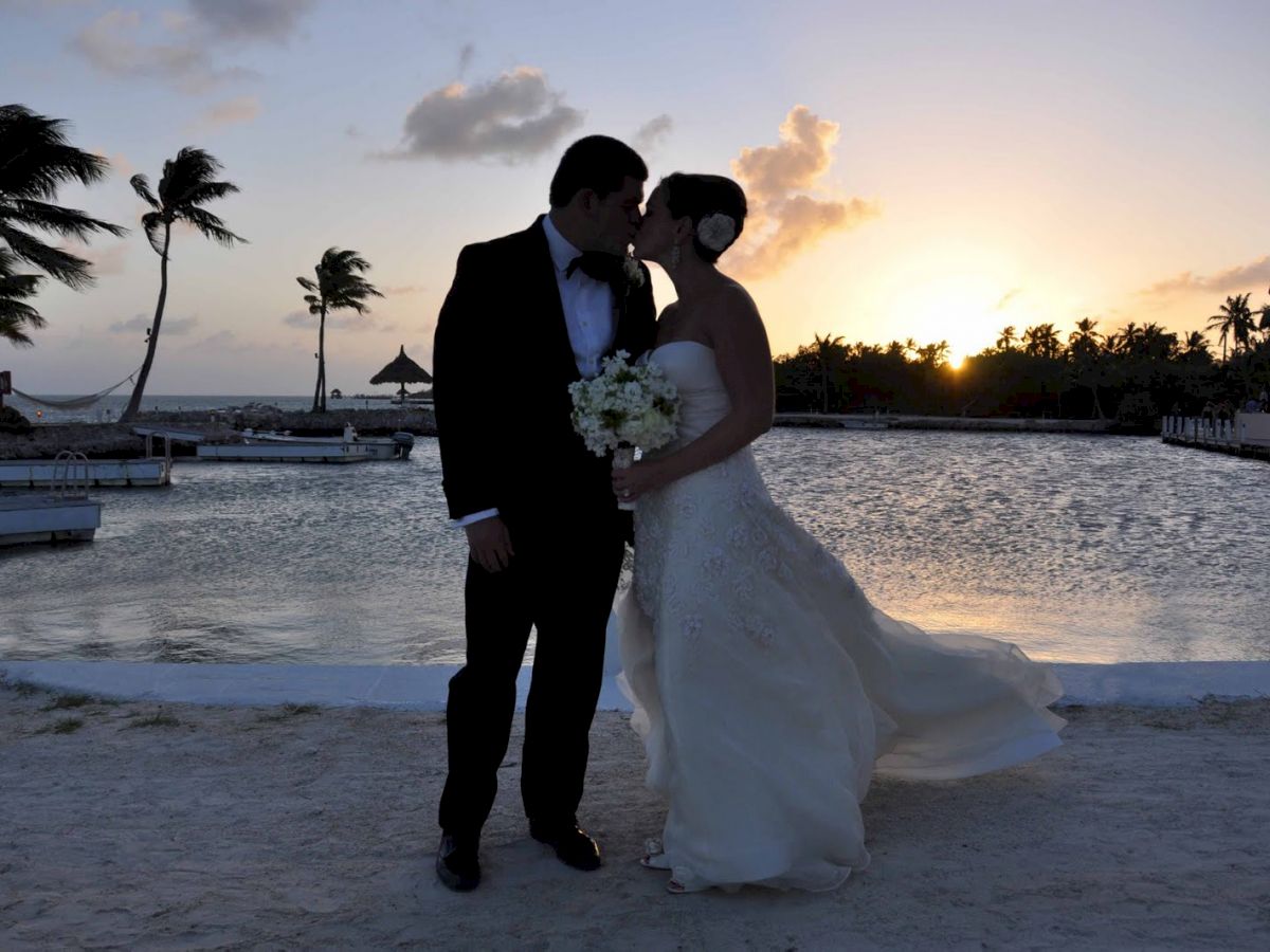 A bride and groom stand close together, facing each other, near a body of water at sunset with palm trees in the background.