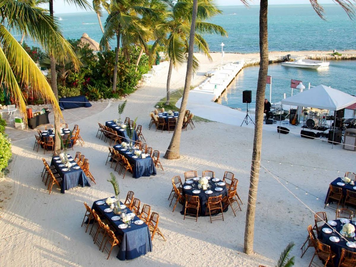 The image shows an outdoor dining setup on a sandy beach with multiple round tables and chairs under palm trees, near the ocean.