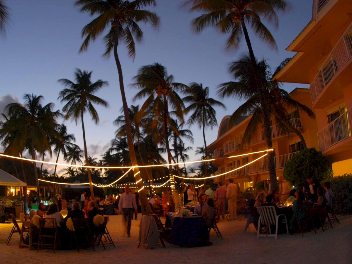 An outdoor evening event with people dining under string lights and palm trees near a beach, with buildings in the background.