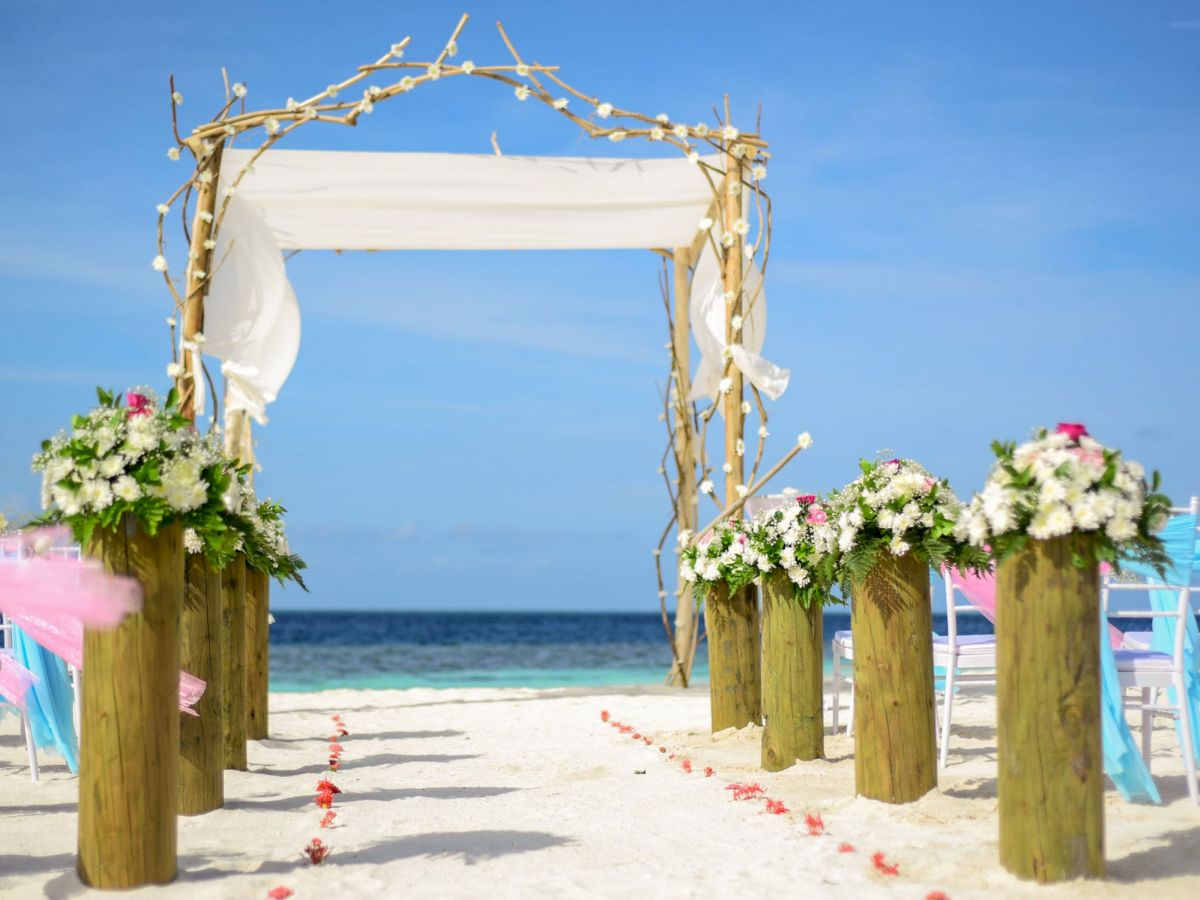 An outdoor beach wedding setup with floral decorations, a wooden arch draped with white fabric, and chairs arranged facing the ocean.