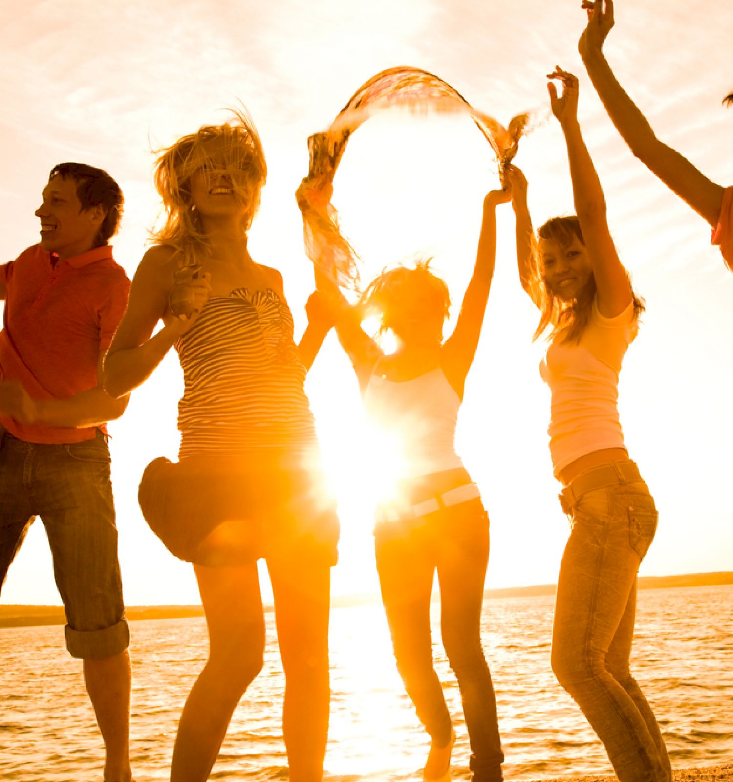 The image shows a group of people joyfully jumping and enjoying themselves on a beach during sunset with the sun shining brightly behind them.