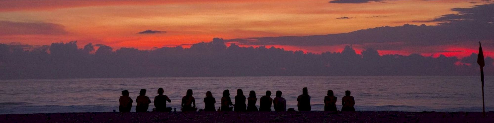 A group of people sitting on a beach, silhouetted against a vibrant sunset sky with clouds and the ocean in the background, creating a serene scene.