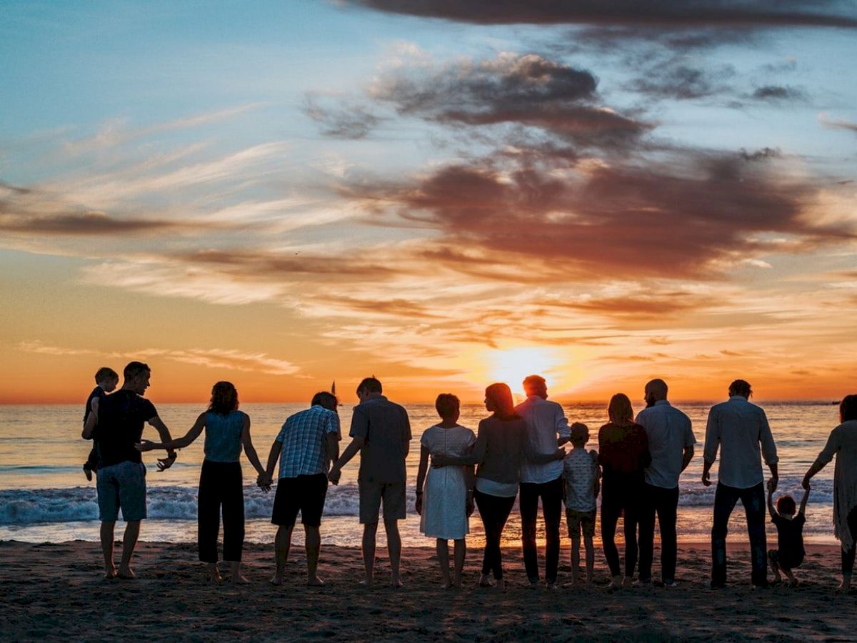 A group of people is standing in a line, holding hands, and facing a sunset on the beach with a picturesque sky filled with clouds ending the sentence.