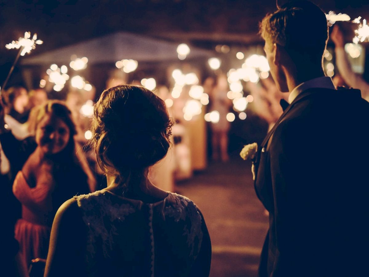 A couple is facing a crowd holding sparklers, likely during a celebratory event or wedding reception at night, creating a warm and festive atmosphere.