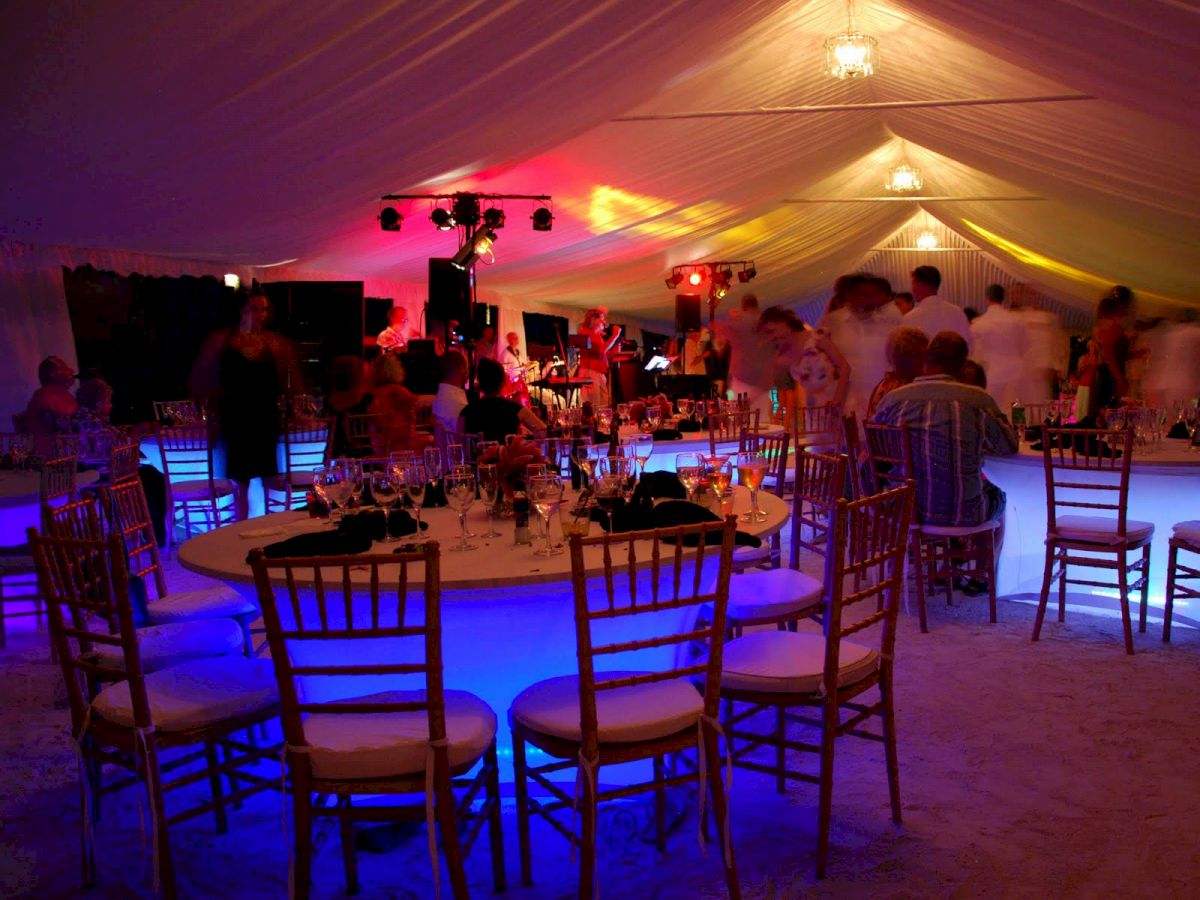 A party or event inside a tent, with vibrant lights and decorated tables and chairs; people are gathered around, enjoying the occasion.