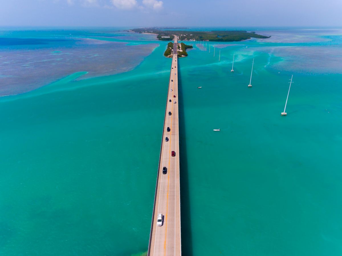 Aerial view of a long bridge over turquoise waters, connecting islands with cars driving along. The sky is clear, and sailboats are docked nearby.