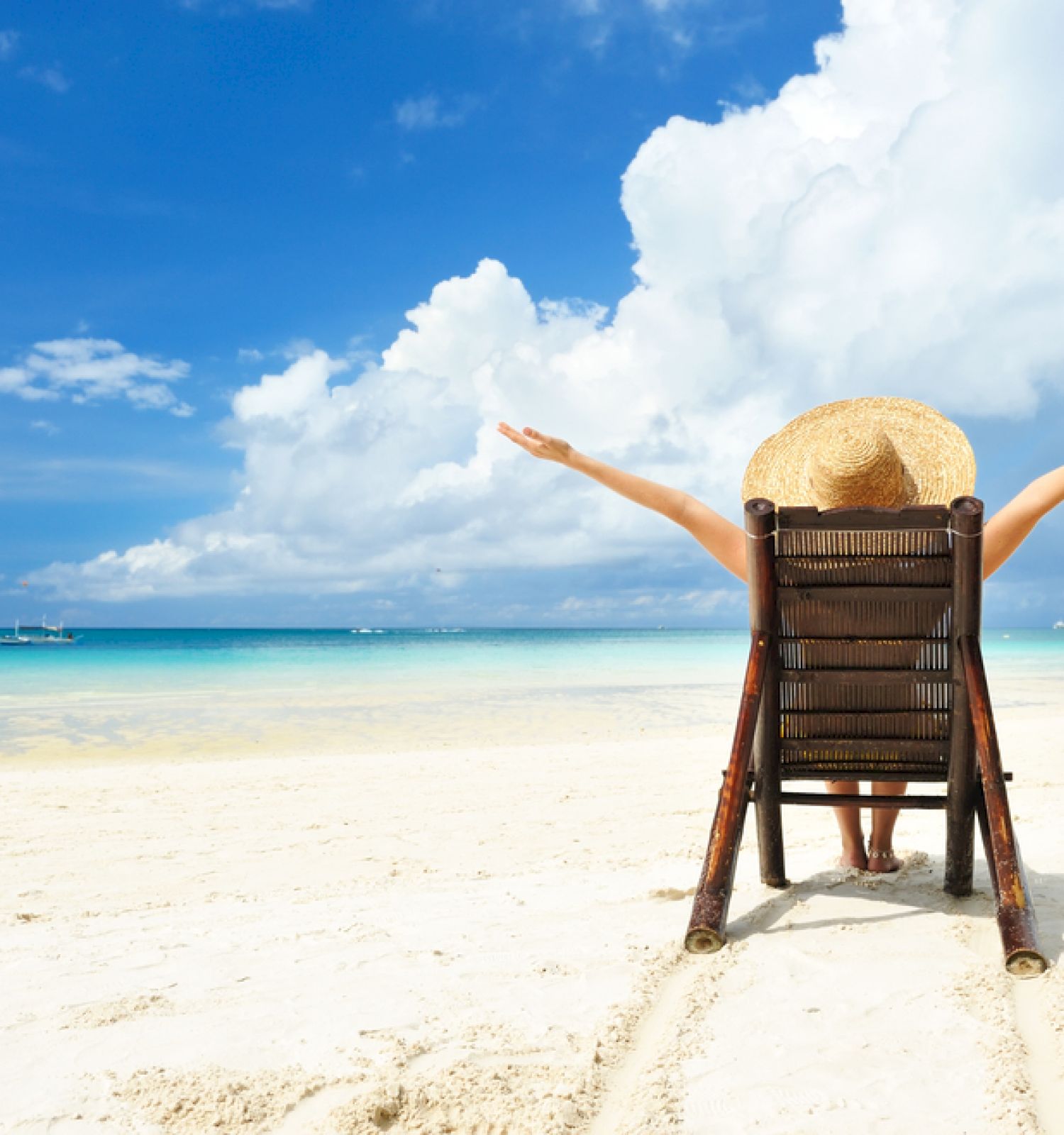 A person sits on a beach chair with arms outstretched, wearing a sunhat, facing the ocean under a bright blue sky with fluffy clouds.