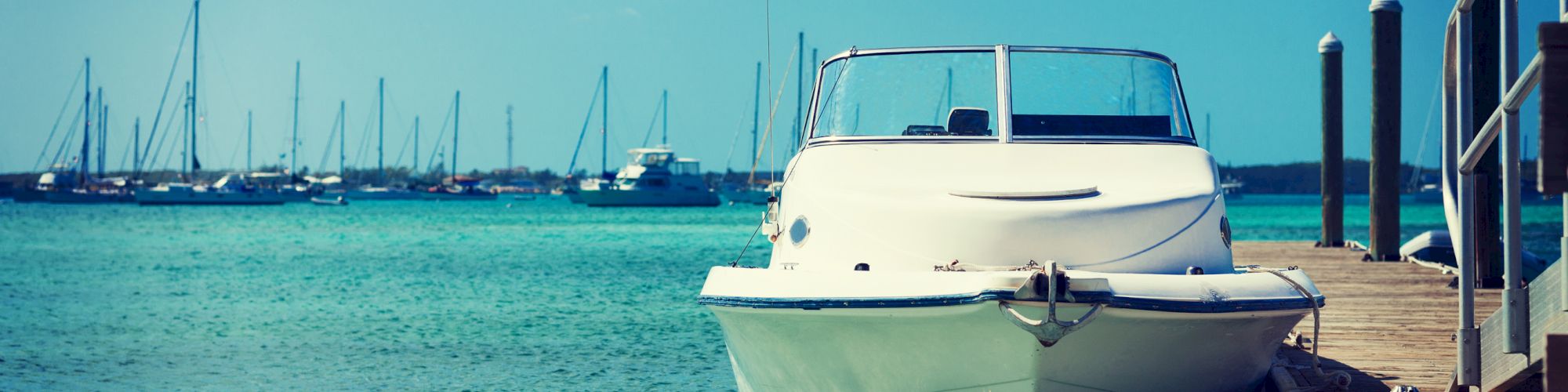 A white boat is docked at a wooden pier on a clear, blue water body, with several other boats in the background under a partly cloudy sky.