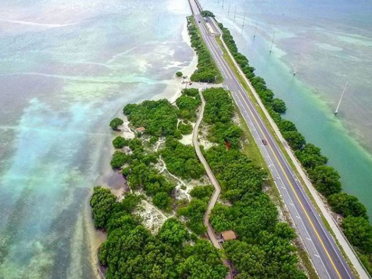 An aerial view of a narrow road stretching across a coastal area, with greenery on both sides and ocean water surrounding it.