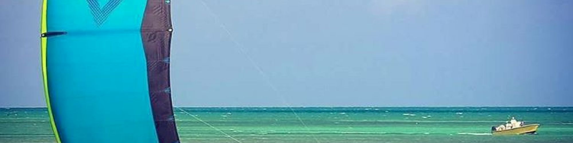 A person is kiteboarding in the ocean with a blue and green kite. The water is clear, and there is a small boat in the background.