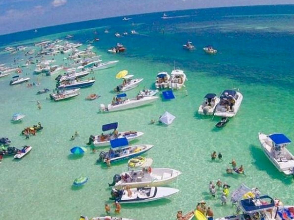 The image shows numerous boats anchored in shallow, clear blue water with people swimming and engaging in various water activities.