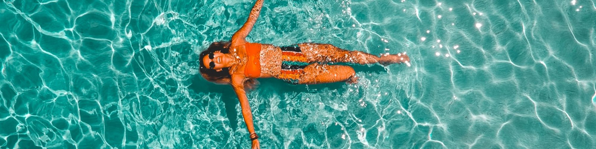 A person is floating on their back in a pool with clear, rippling water, wearing an orange swimsuit.