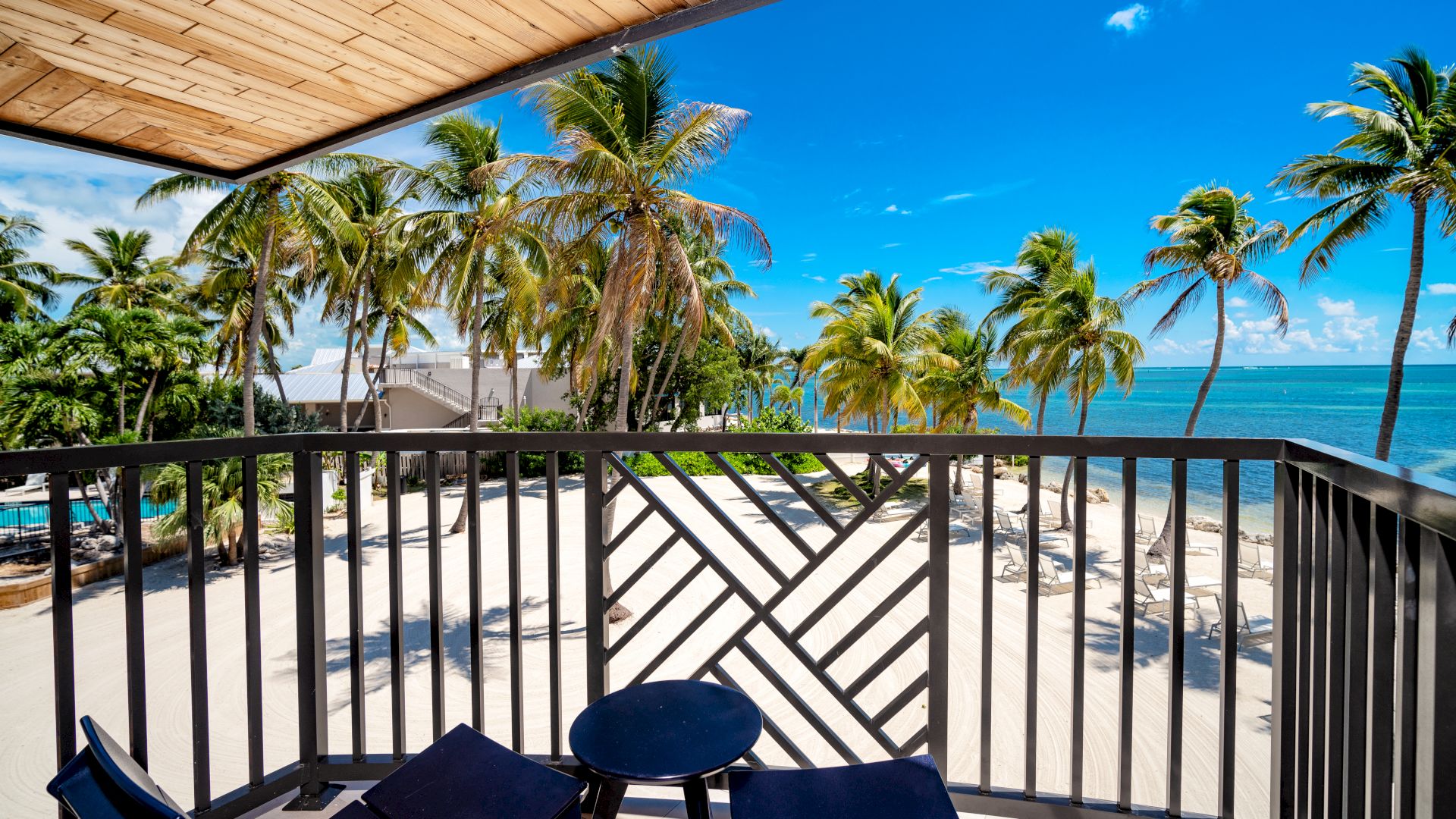 A beachfront balcony with two chairs and a table overlooks a sandy beach lined with palm trees under a clear blue sky.