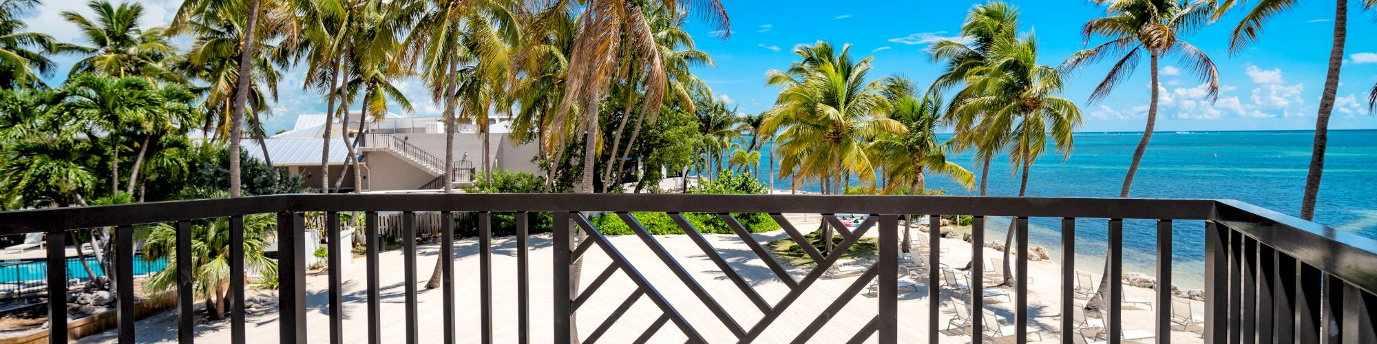 A beachfront balcony with two chairs and a table overlooks a sandy beach lined with palm trees under a clear blue sky.