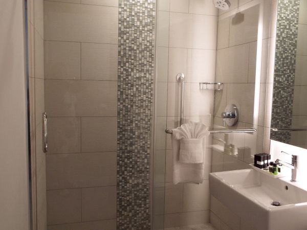 A modern bathroom with a glass walk-in shower, rainfall showerhead, tiled walls, and a sink with folded towels.