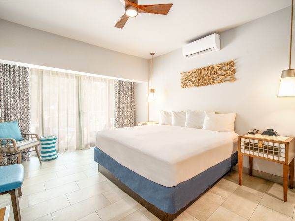 A modern hotel room with a king-size bed, bedside table, seating area, large window, air conditioning, and minimalist decor.