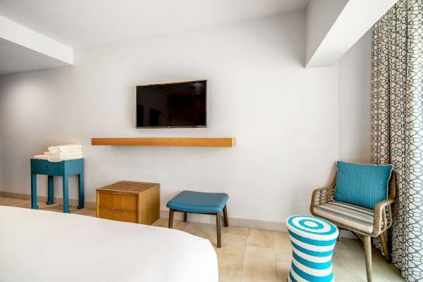 A minimalist hotel room with a wall-mounted TV, small table with towels, stool, chair with pillow, striped ottoman, and patterned curtains.