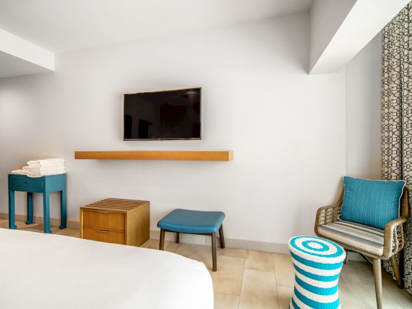 A minimalist hotel room with a wall-mounted TV, small table with towels, stool, chair with pillow, striped ottoman, and patterned curtains.