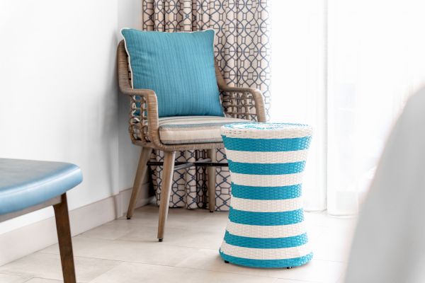 A corner with a wicker chair, a blue cushion, and a small round striped stool in blue and white. The backdrop features a patterned curtain.