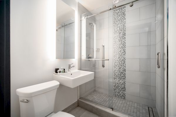 This image shows a modern bathroom with a toilet, a sink with a mirror, and a shower with a sliding glass door and tiled walls.