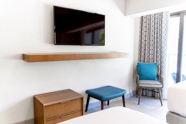 The image shows a modern room with a wall-mounted TV, floating shelf, small table, chair, and a window with patterned curtains.