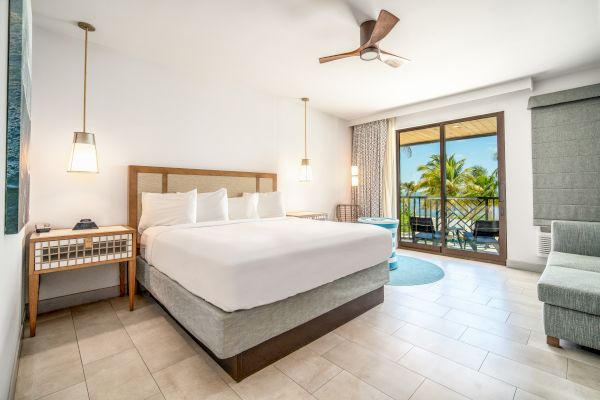 A hotel room with a king-size bed, bedside tables, a ceiling fan, and a balcony overlooking a tropical view.