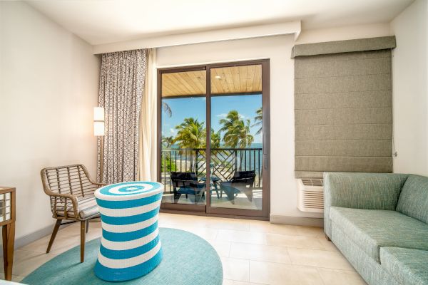 A cozy, sunlit room with modern furniture, featuring a balcony overlooking a tropical scene with palm trees and a blue sky.