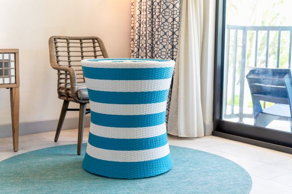 A blue and white striped wicker stool on a round blue rug, next to a wicker chair in a room with floor-length curtains and a sliding glass door.