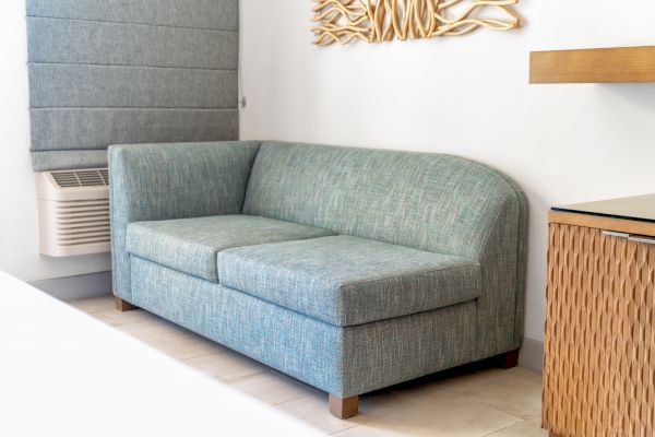 A blue-green couch is set in a bright room with a wall-mounted air conditioning unit, decorative wall art, and a wicker cabinet nearby, ending the sentence.