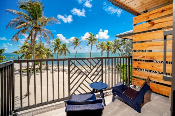 A balcony with two chairs and a small table, overlooking a beautiful beach with palm trees and a clear blue sky in the background.