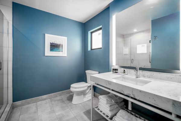 A modern bathroom with a blue accent wall, a white sink on a marble countertop, a toilet, stacked towels, a shower, and a framed picture.