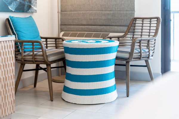 The image shows two wicker chairs with blue cushions and a striped blue and white cylindrical table between them in a cozy, modern room.