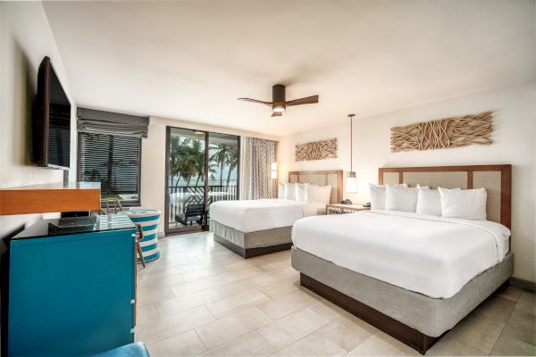 A modern hotel room with two white beds, a flat-screen TV, a desk, a blue mini-fridge, and a balcony with a view of palm trees is pictured.