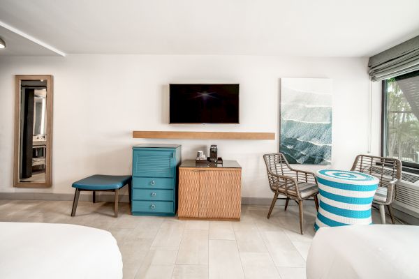 A modern hotel room with a flat-screen TV, a wooden cabinet, a blue chest of drawers, chairs, a striped table, and a large window.