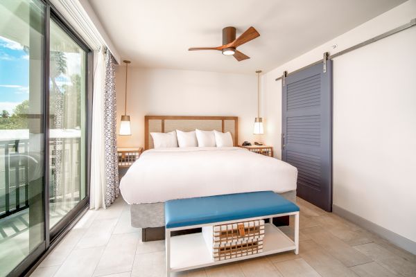 A modern hotel room with a large bed, bedside lamps, a ceiling fan, a blue sliding door, a bench, and a balcony with a view of the outdoors.