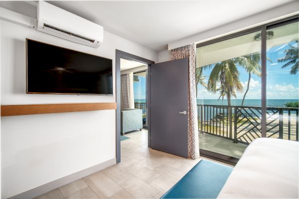 A bright room with a TV on the wall, open door leading to a balcony with a sea view and palm trees, and a glimpse of a bed in the corner.