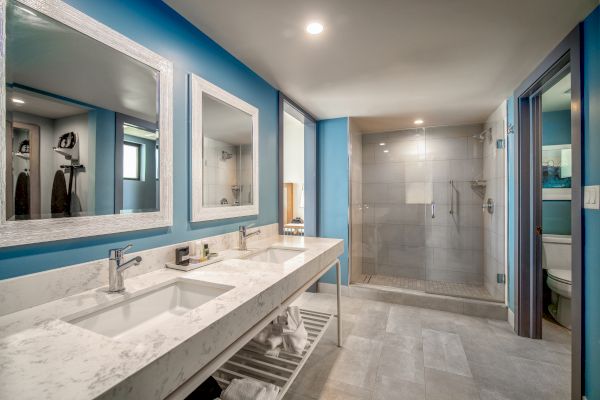 A modern bathroom with a double sink vanity, large mirrors, blue walls, a walk-in shower, and a separate toilet area.