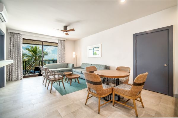 A modern living room features a gray couch, wicker chairs, a round dining table, a ceiling fan, large windows with curtains, and a view of palm trees outside.