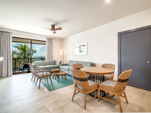 A modern living room features a gray couch, wicker chairs, a round dining table, a ceiling fan, large windows with curtains, and a view of palm trees outside.