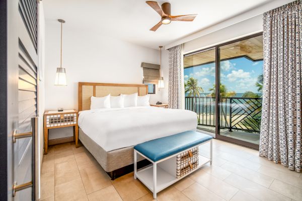 A bright, modern bedroom with a king-sized bed, ceiling fan, bedside tables, and a balcony overlooking a scenic beach view through sliding glass doors.