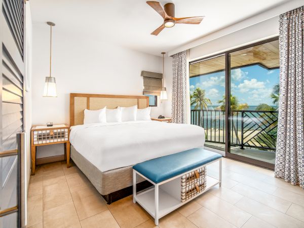 A bright, modern bedroom with a king-sized bed, ceiling fan, bedside tables, and a balcony overlooking a scenic beach view through sliding glass doors.