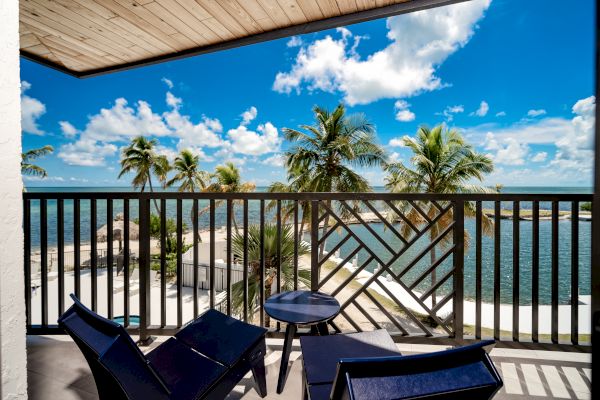 A balcony with two chairs and a small table, overlooking a tropical beach with palm trees and clear blue skies.